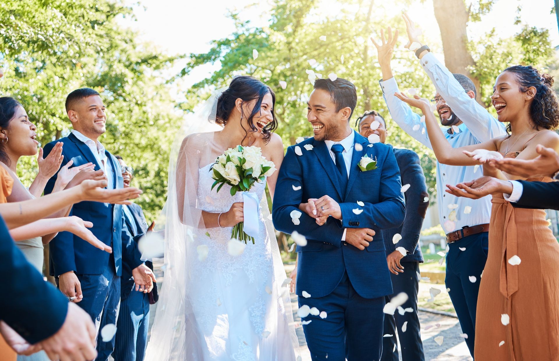 Bride and groom smiling and walking as guests toss flower petals at outdoor wedding.