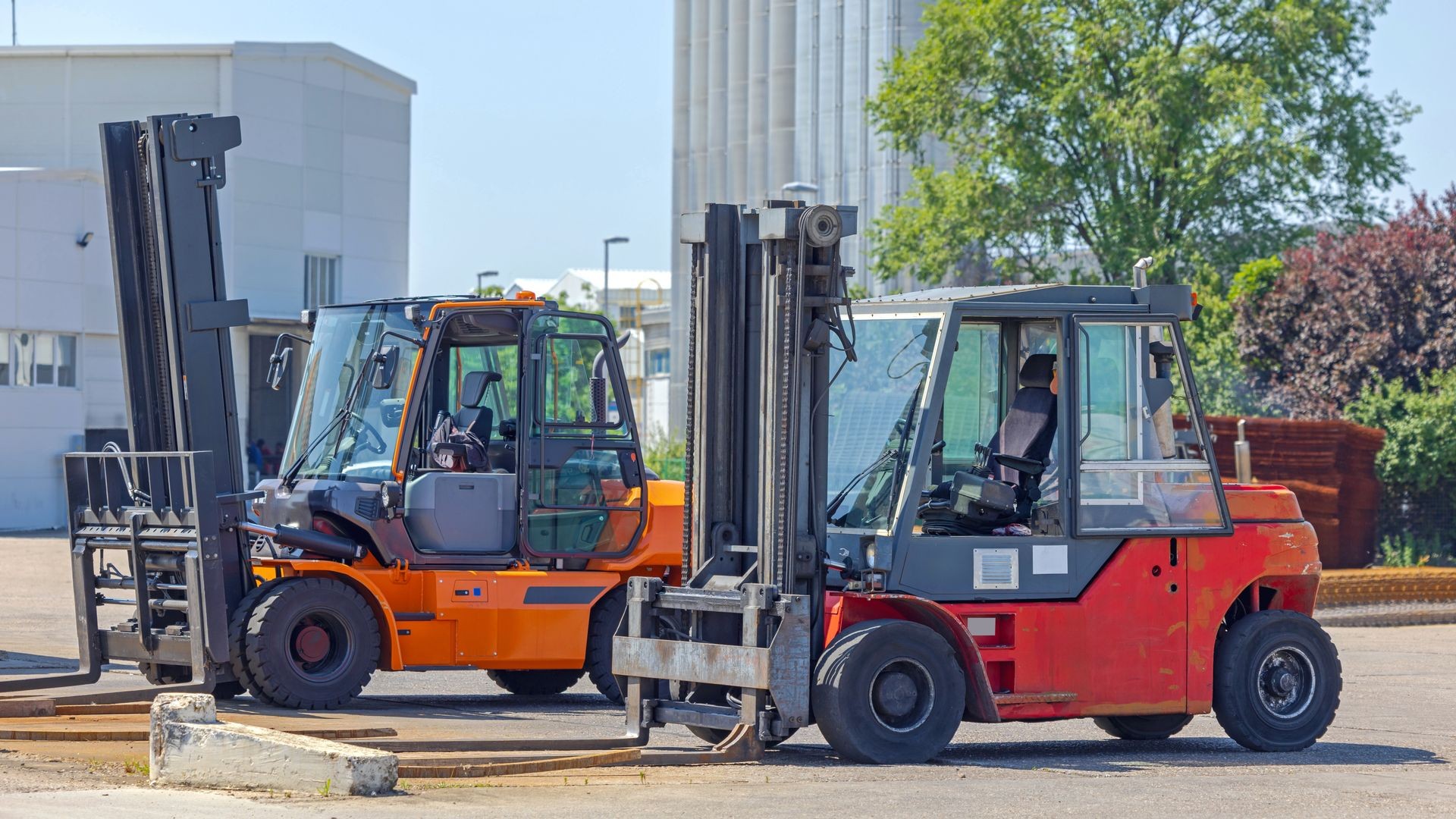 Two forklifts parked outdoors in an industrial area with buildings in the background and trees providing shade.