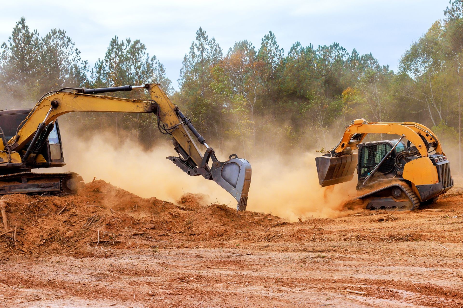 Excavator and skid steer loader moving dirt on a construction site with trees in the background.