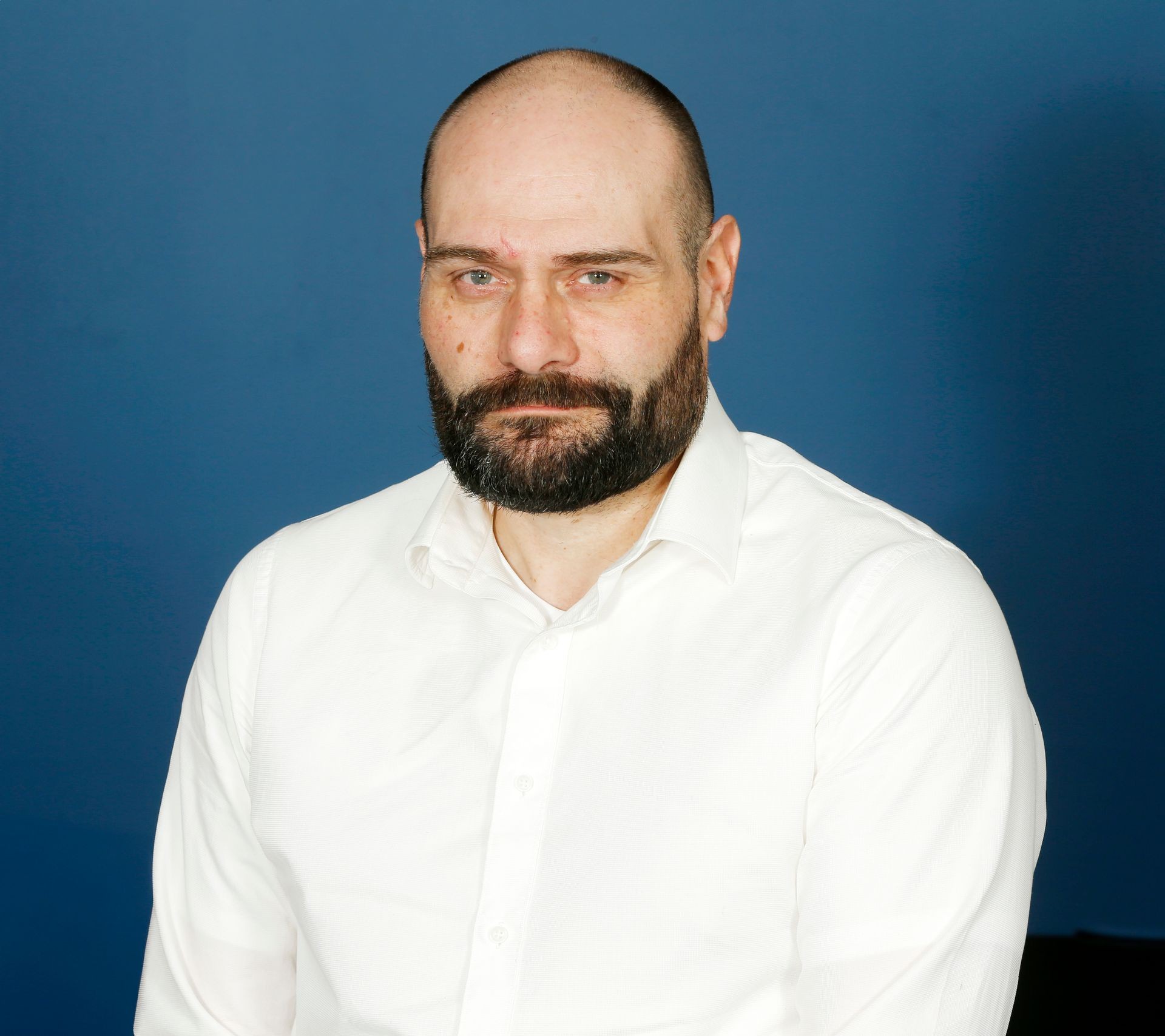 Man with a beard and white shirt standing against a blue background.