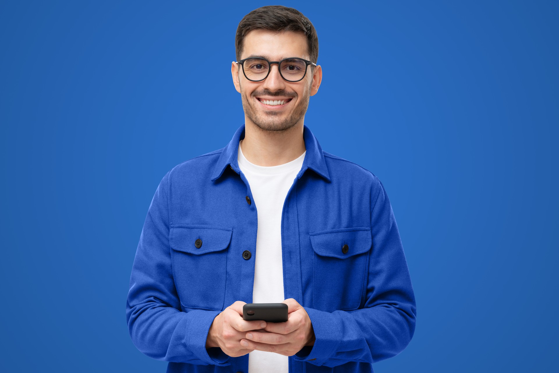 Young man in casual blue shirt and glasses, holding phone, looking at camera with smile